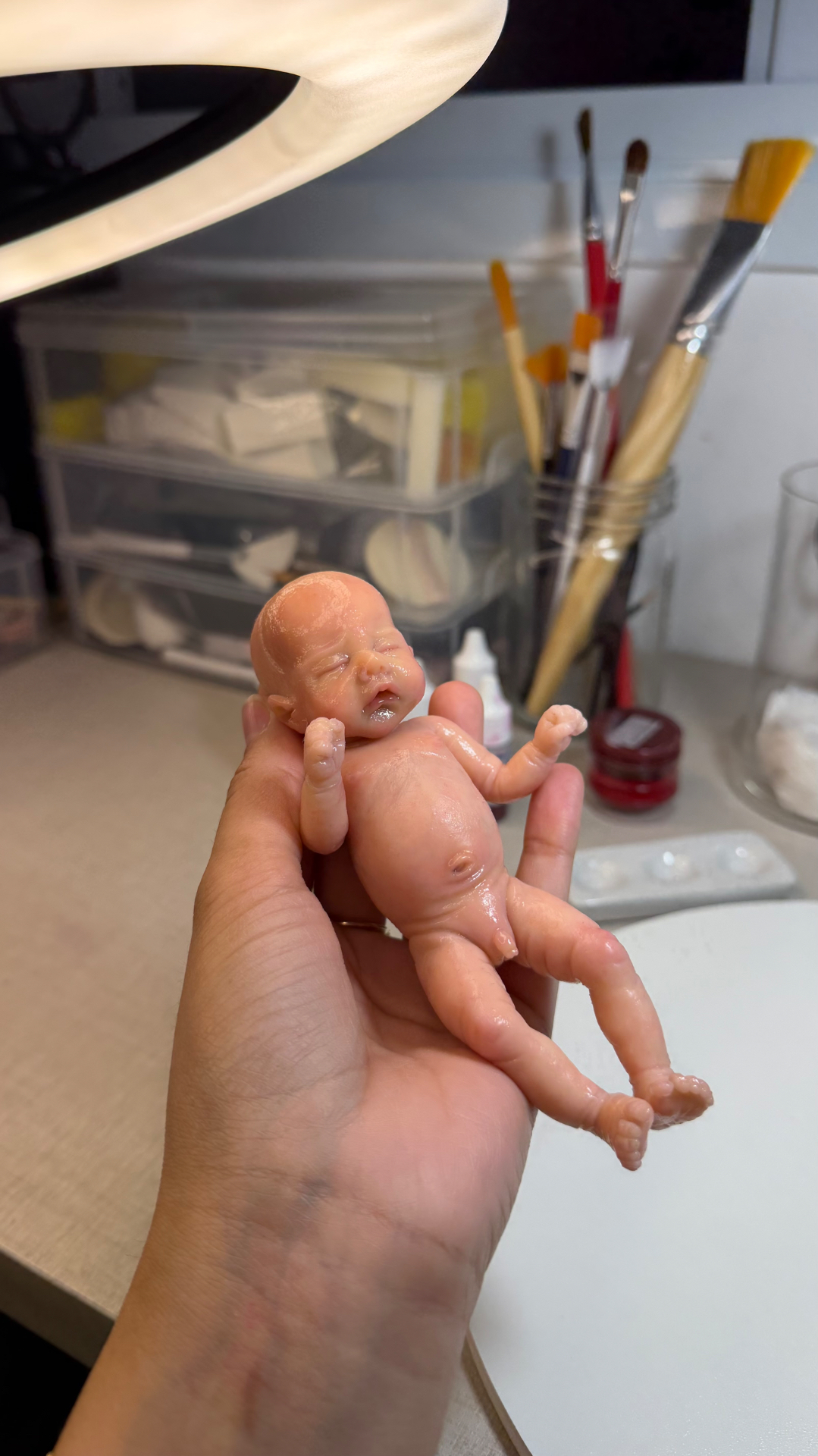 A miniature silicone baby resting in the artist's palm beside her brushes